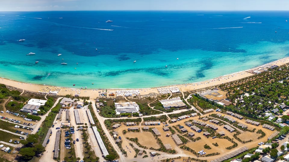 An aerial view of a coastal town with a curved bay, clear turquoise waters, boats, and a densely populated area with buildings and roads.