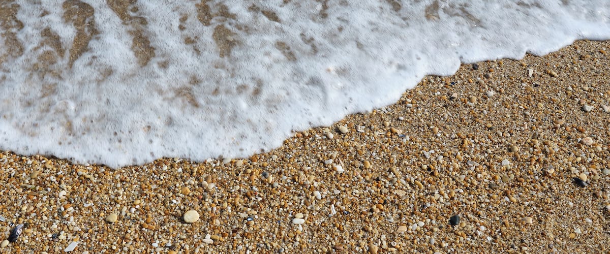 A sandy beach with small pebbles and a foamy wave.