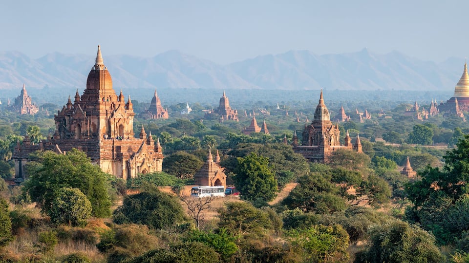 A landscape with numerous ancient temples and a prominent golden stupa amidst lush greenery and mountains in the background.