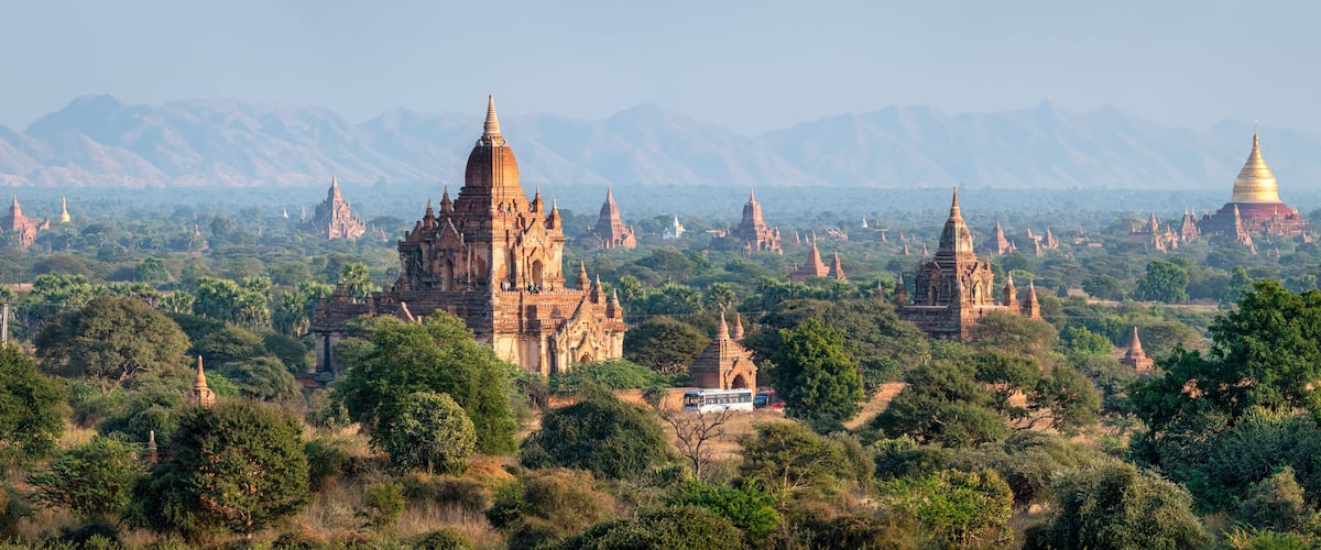 A landscape with numerous ancient temples and a prominent golden stupa amidst lush greenery and mountains in the background.