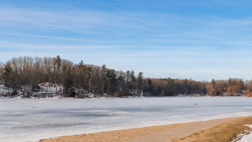 A frozen lake with a sandy beach in the foreground and a forested area in the background.
