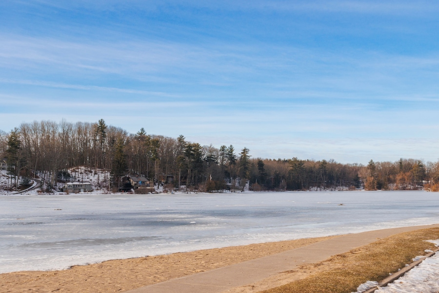 A frozen lake with a sandy beach in the foreground and a forested area in the background.