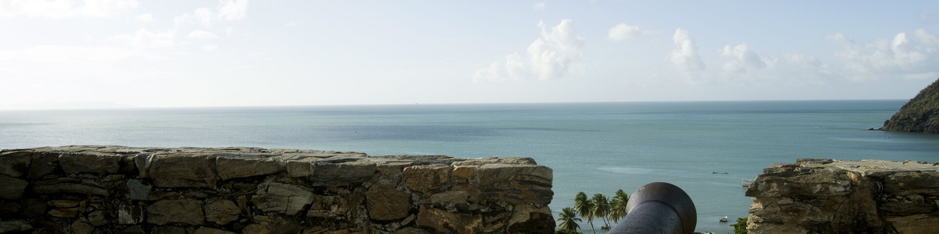 A cannon overlooking a coastal view with a stone wall and a clear sky.