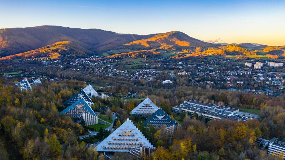 Eine Stadt liegt in einem Tal, mit Bergen im Hintergrund, umgeben von herbstlich gefärbten Bäumen.