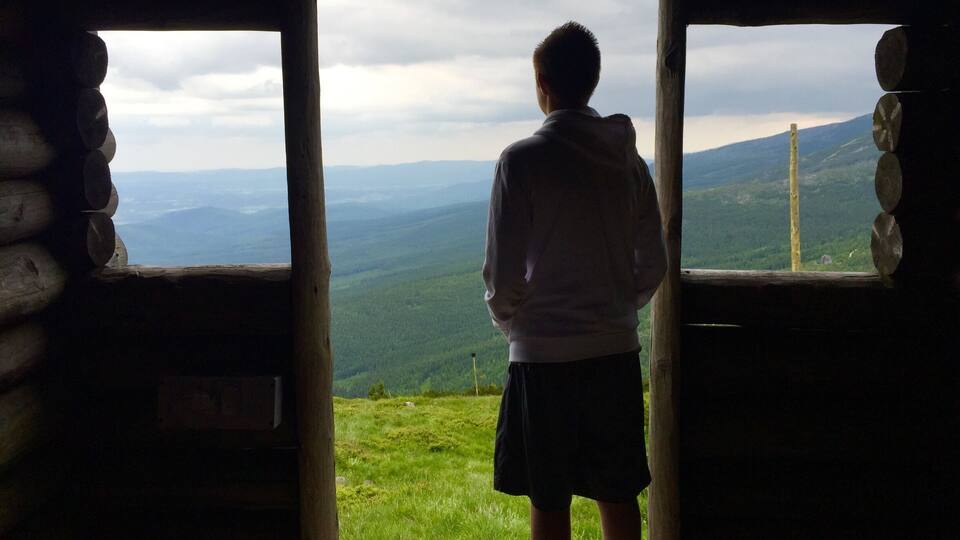 A person standing at a wooden doorway looking out at a mountainous landscape.