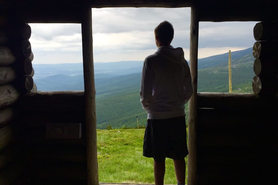 A person standing at a wooden doorway looking out at a mountainous landscape.