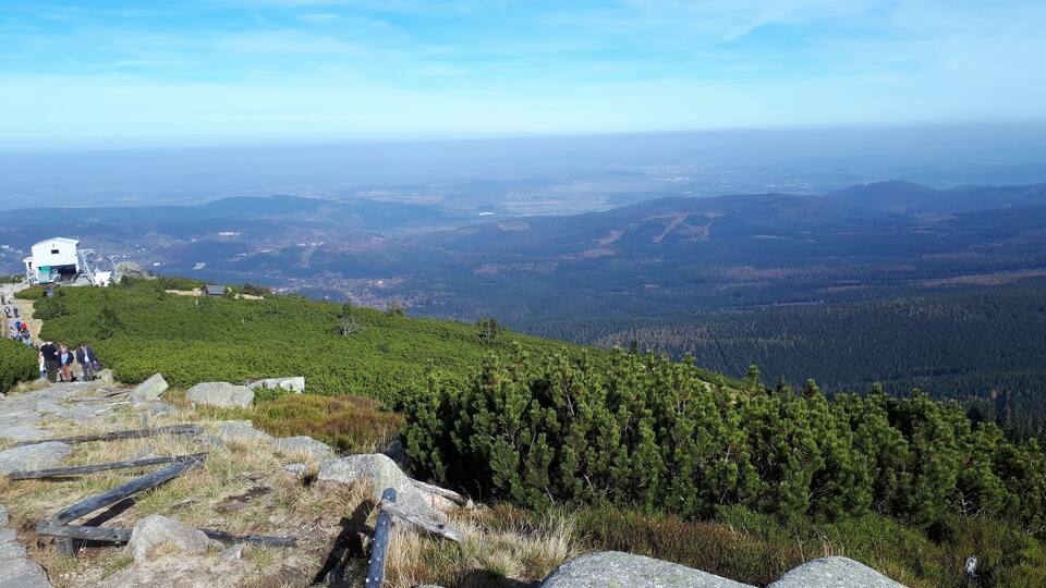 A mountain trail with a wooden railing, rocky terrain, and a distant view of a valley with a cabin.