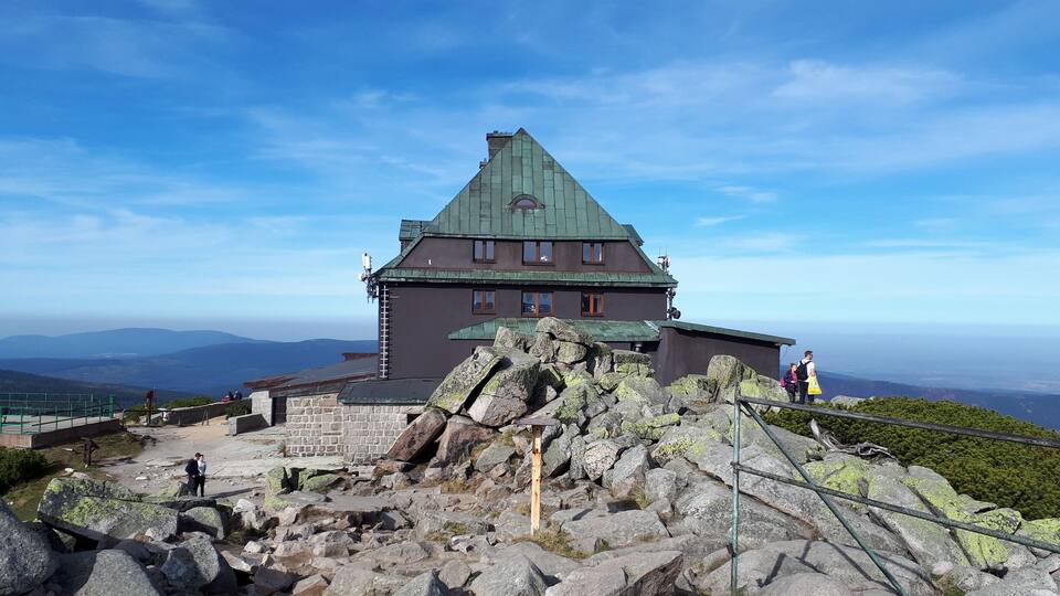 A large stone building with a green roof, surrounded by rocky terrain and a clear blue sky.