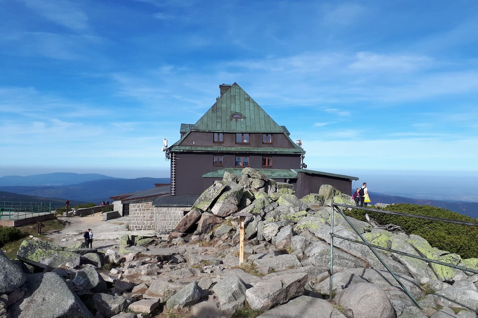 A large stone building with a green roof, surrounded by rocky terrain and a clear blue sky.