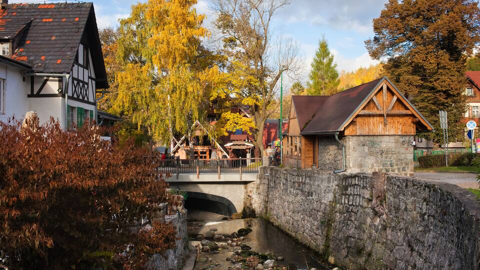 A stone bridge over a narrow stream, surrounded by autumn-colored trees and traditional buildings.