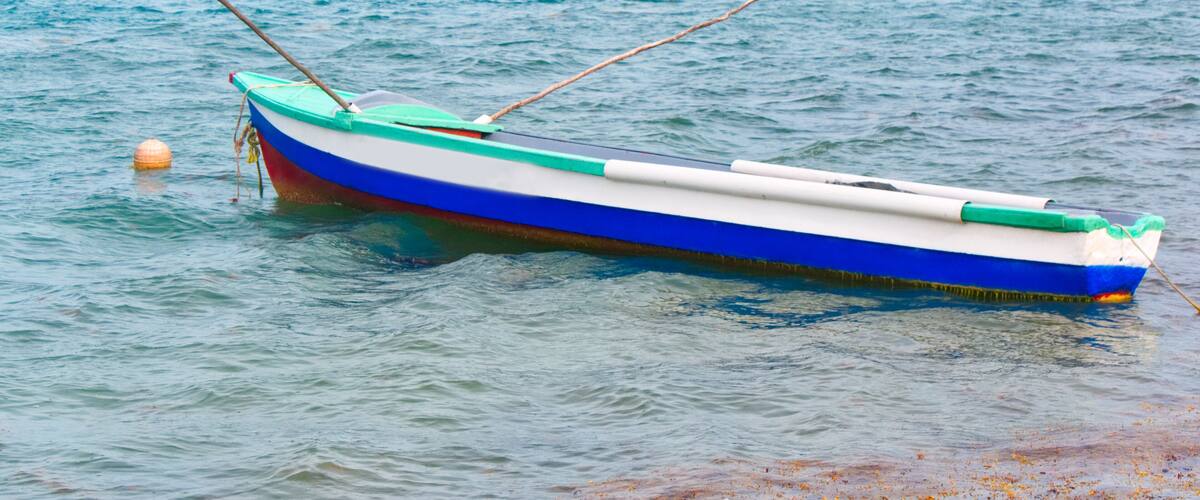 A small boat with a green and blue hull, anchored near a rocky shoreline with green hills in the background.