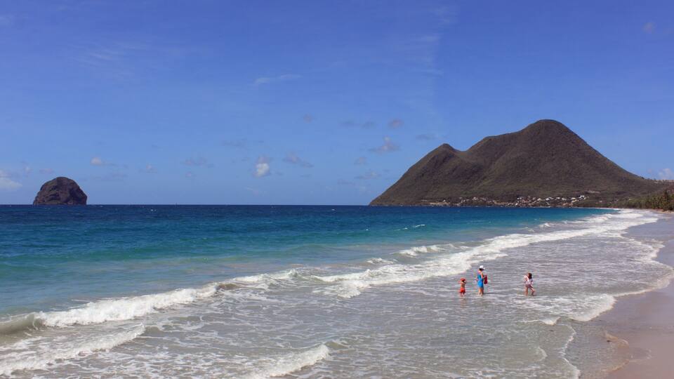 A beach with clear blue water, a rocky outcrop, and a mountain in the background.