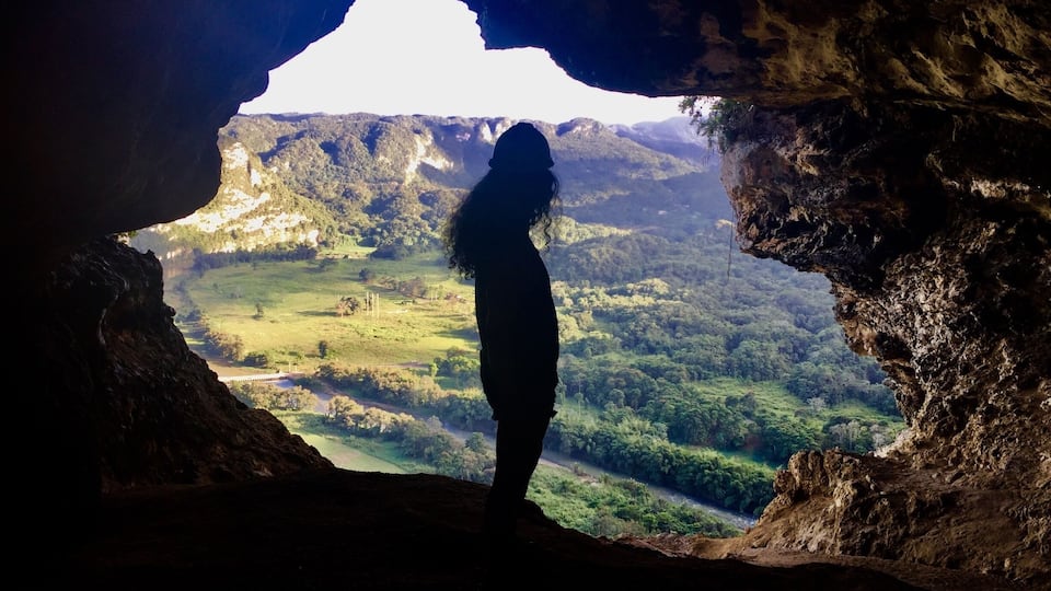 A person silhouetted against a cave opening, overlooking a lush valley with a river and distant mountains.