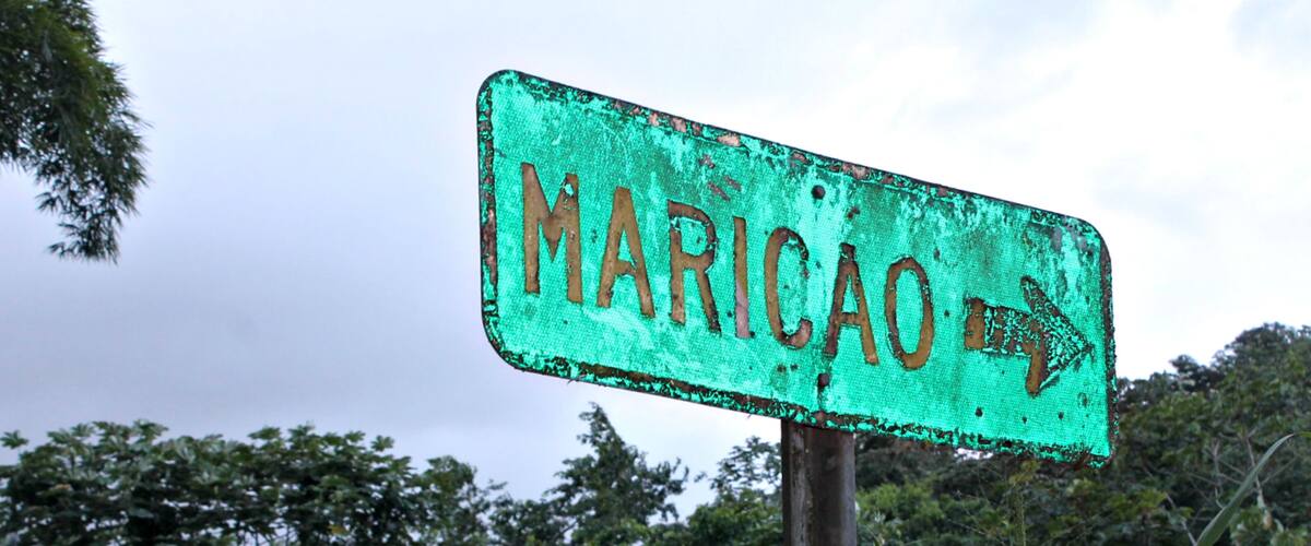 A weathered green sign with the word 'MARICAO' and an arrow pointing right, surrounded by lush greenery.