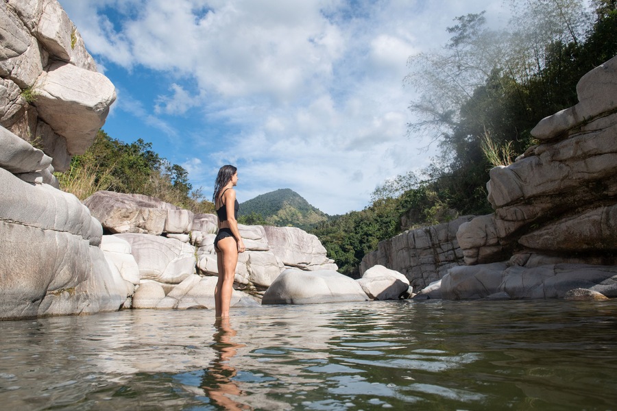A woman standing in a rocky river with large boulders and a mountain in the background.