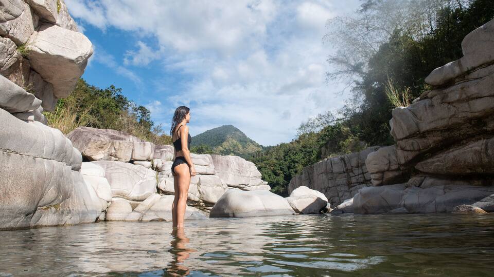 A woman standing in a rocky river with large boulders and a mountain in the background.