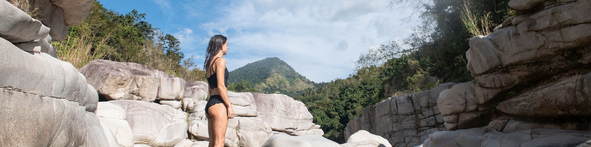 A woman standing in a rocky river with large boulders and a mountain in the background.