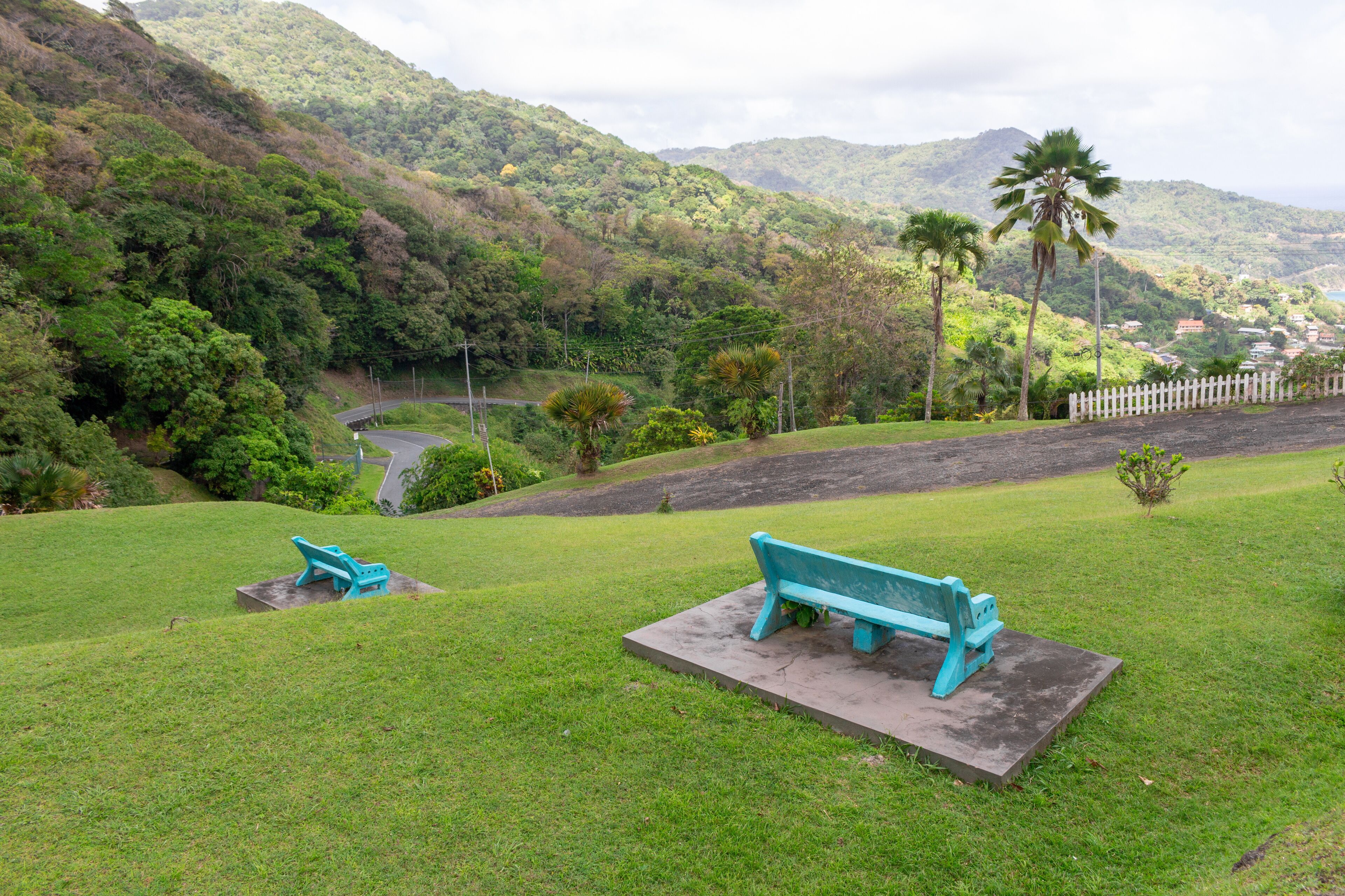 A park with two turquoise benches, lush green grass, and a white picket fence.