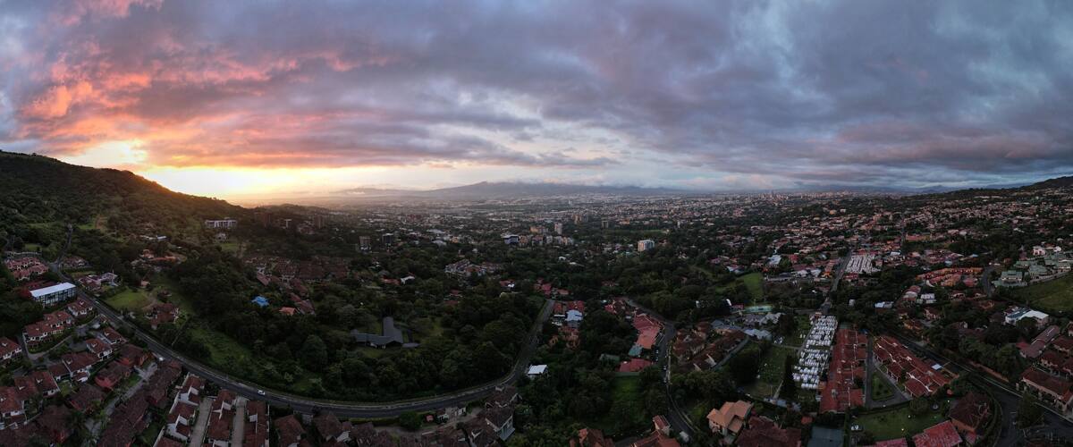 A panoramic view of a city with a winding road, dense residential areas, and a backdrop of hills under a dramatic sunset sky.