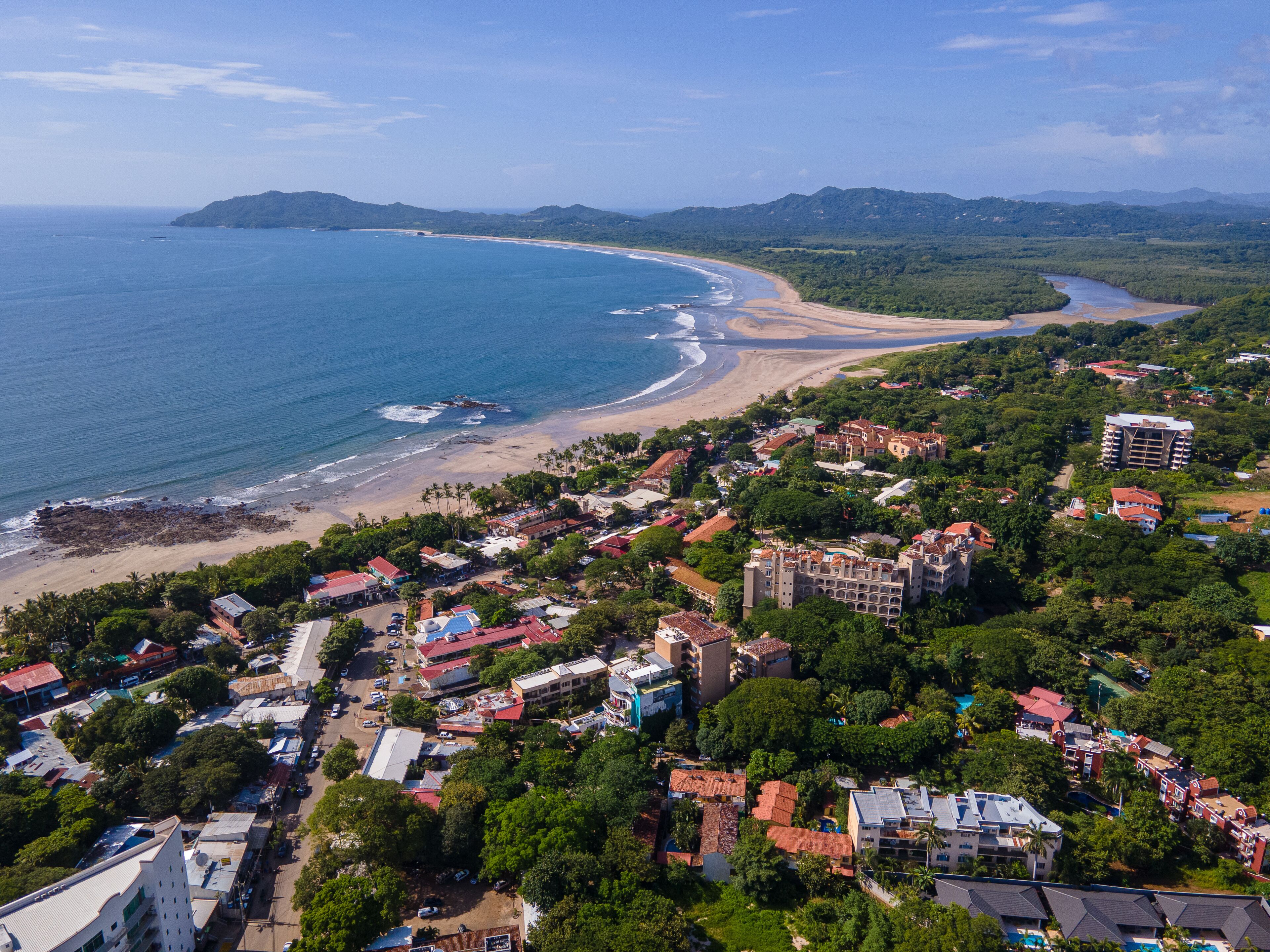 A coastal town with a beach, dense greenery, and a mix of residential and commercial buildings.