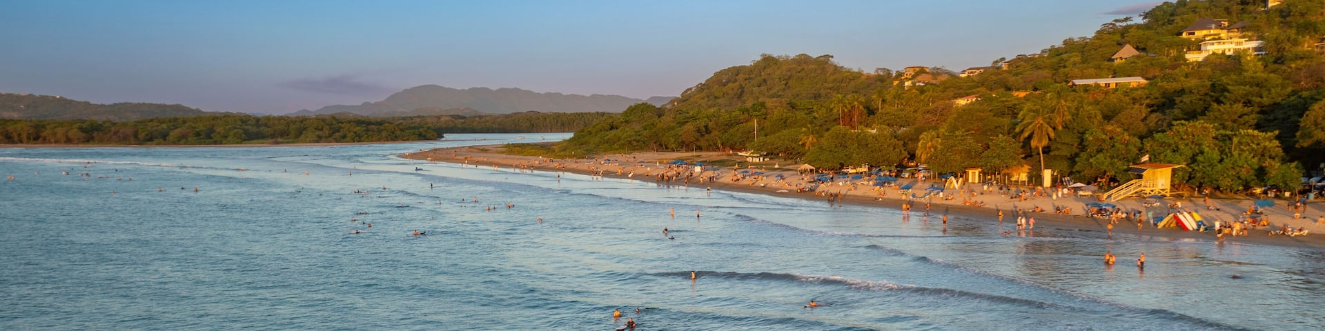 A coastal beach with surfers and swimmers, a clear sky, and a hilly landscape with buildings.