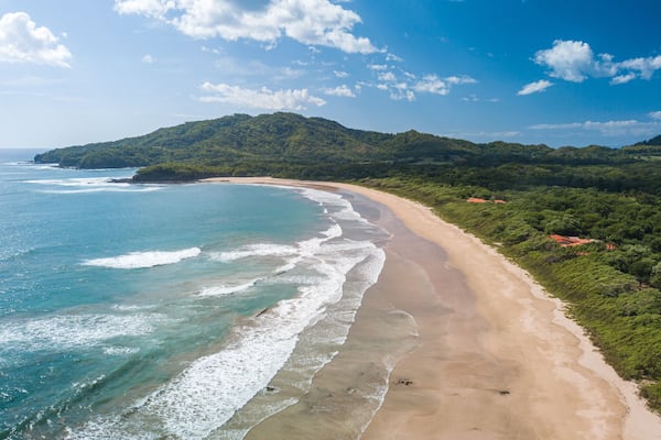 Una playa con aguas azules cristalinas, una orilla de arena y colinas verdes al fondo.