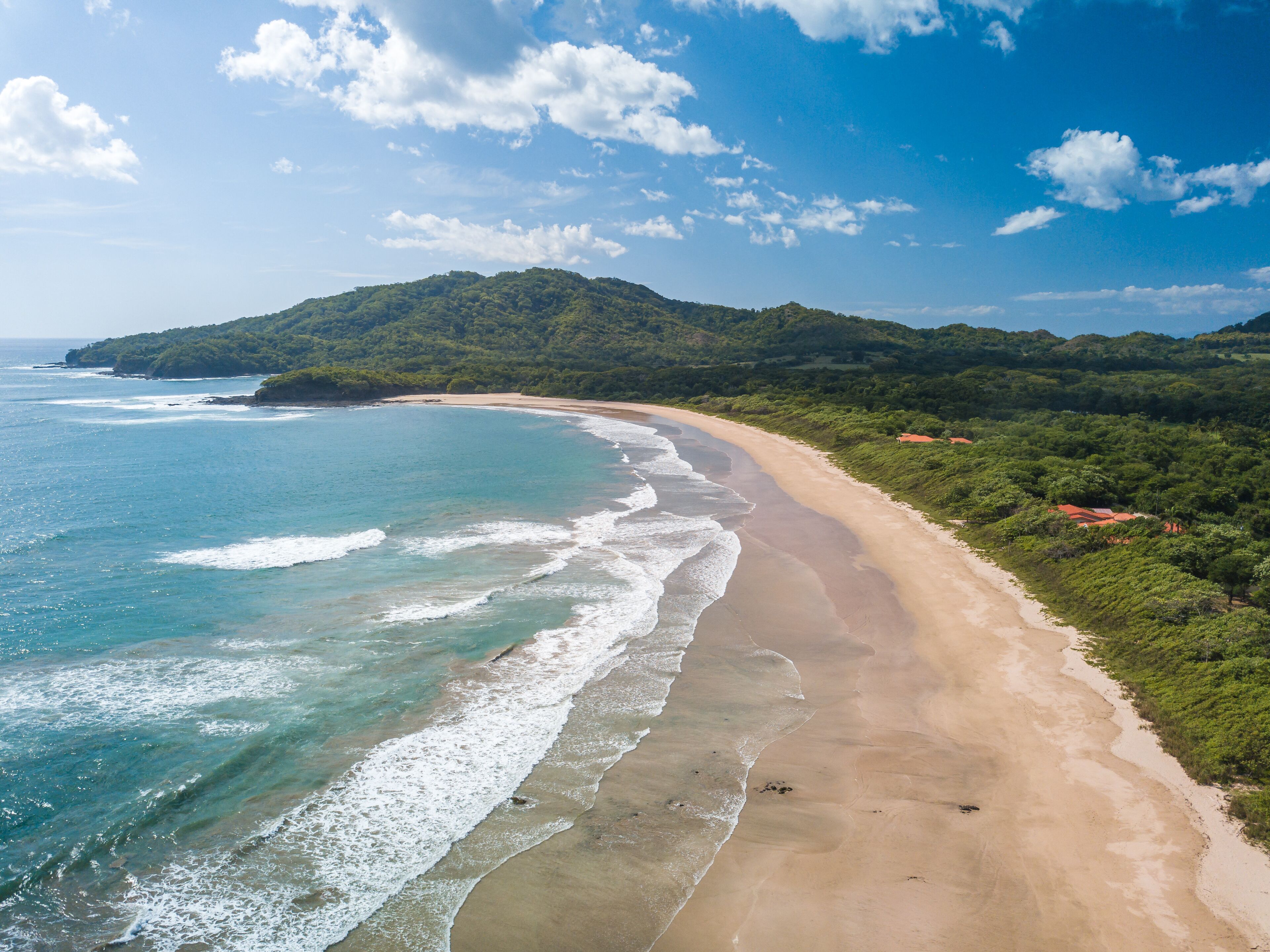 A beach with clear blue waters, a sandy shore, and green hills in the background.