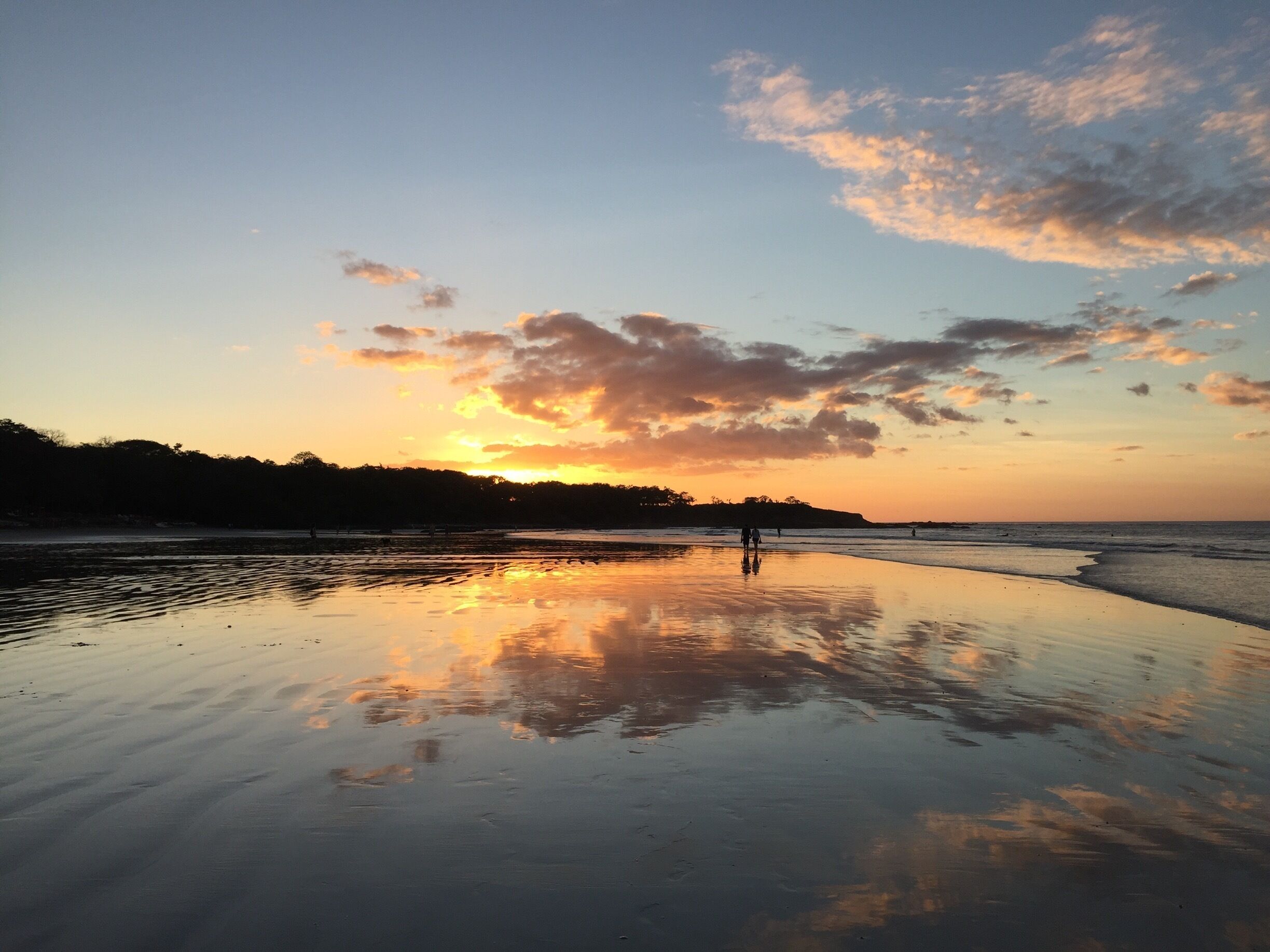 A beach at sunset with a calm sea, a person walking, and a reflection of the sky on the wet sand.
