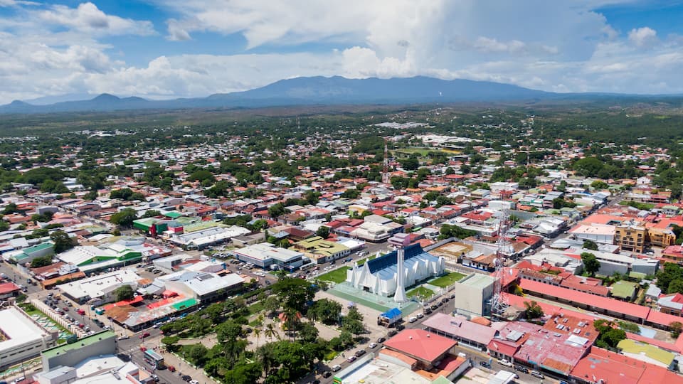 An aerial view of a city with a mix of residential and commercial buildings, a prominent white church, and a mountain range in the background.