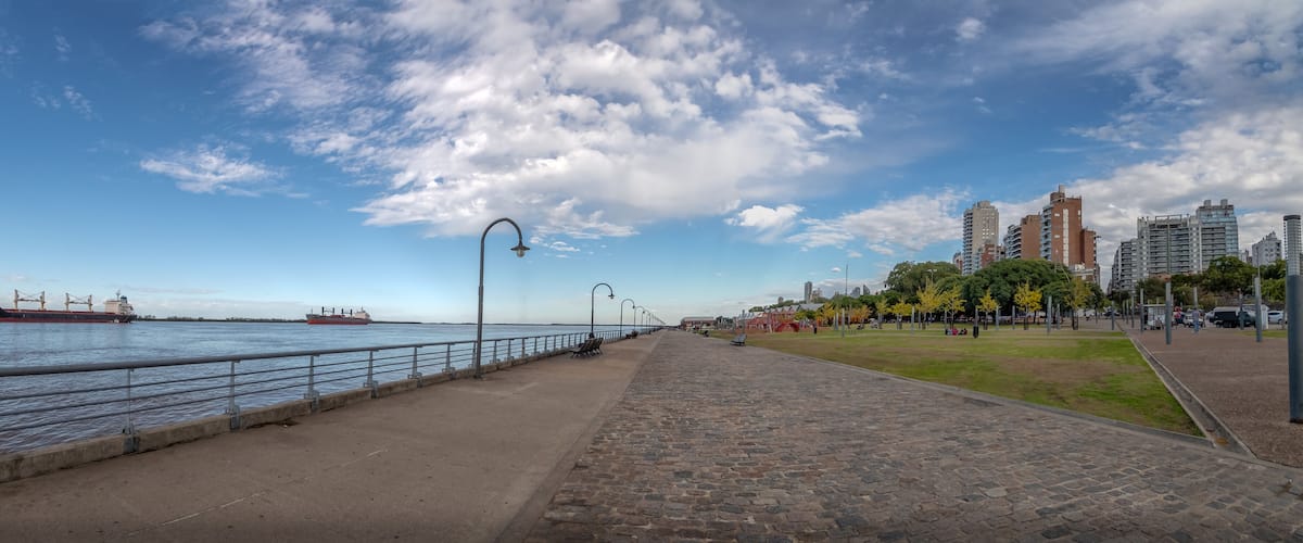A waterfront promenade with a large cargo ship, a clear sky, and modern buildings in the background.