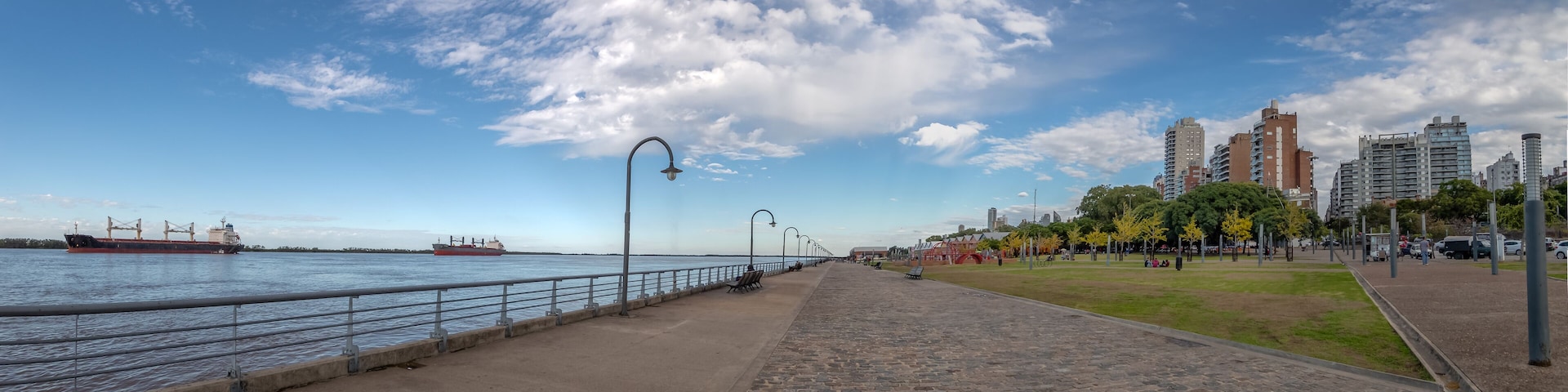 A waterfront promenade with a large cargo ship, a clear sky, and modern buildings in the background.