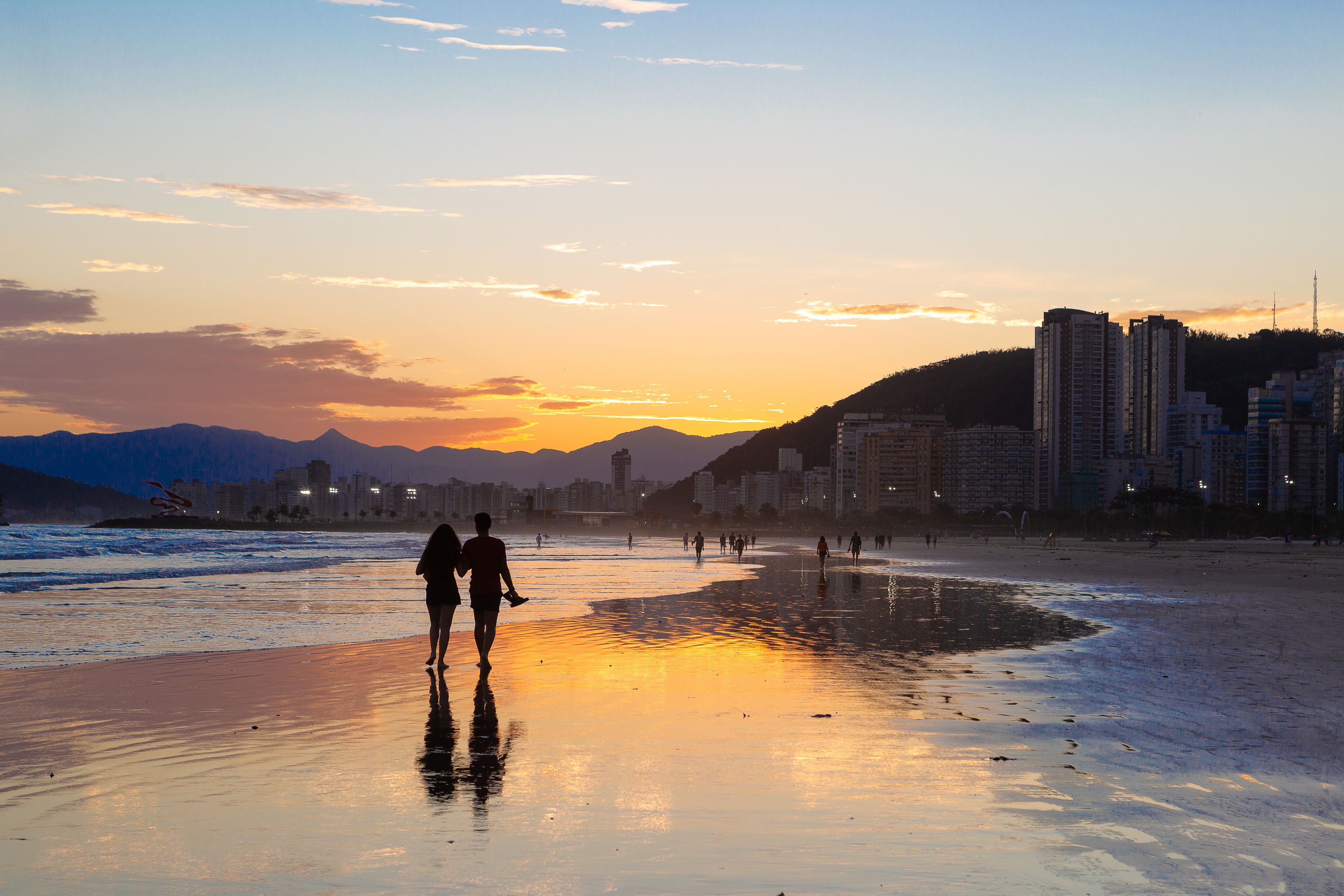 A beach at sunset with silhouettes of people walking, a cityscape in the background, and mountains in the distance.
