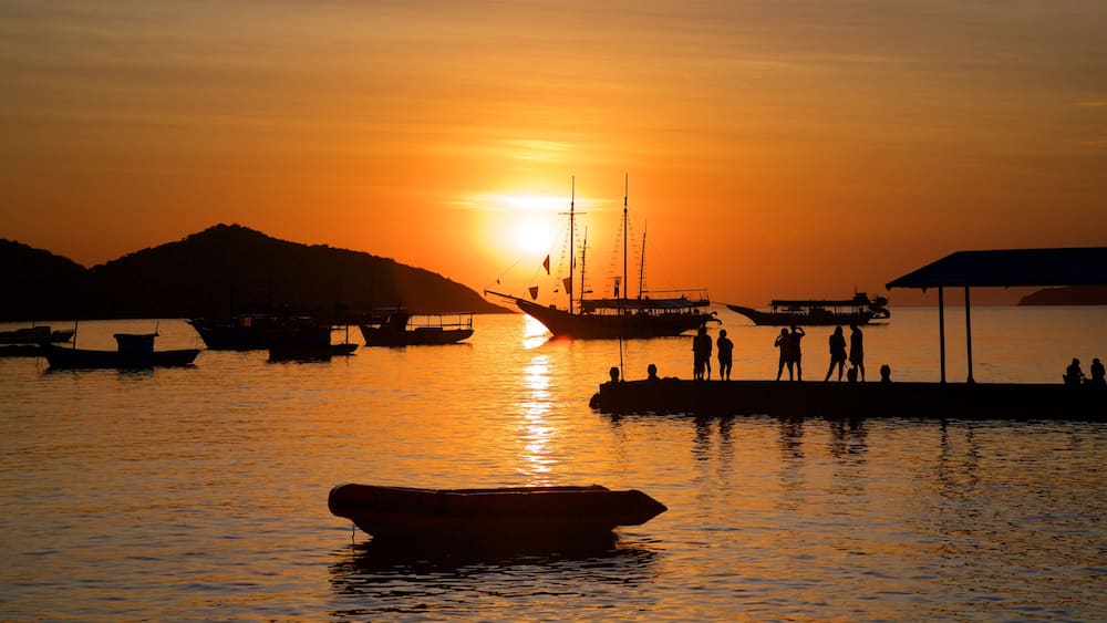 Un coucher de soleil sur un port, avec des bateaux et des personnes se détachant en silhouette sur le ciel.