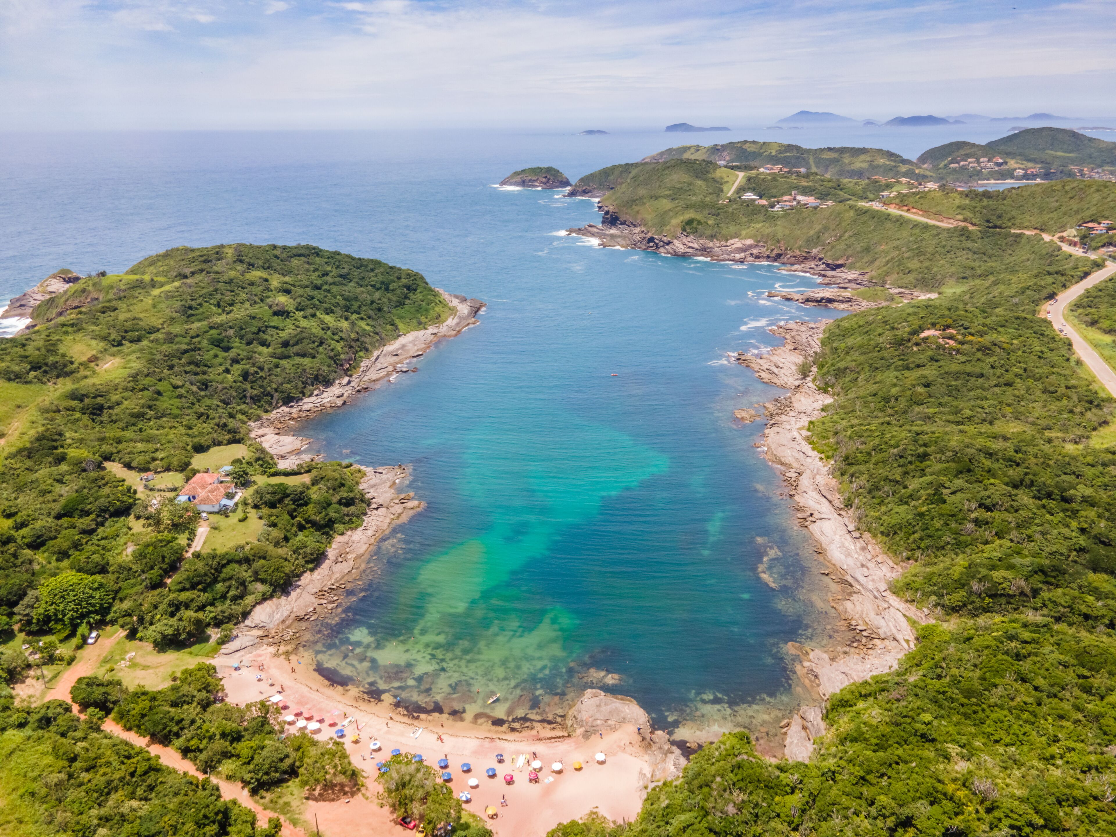 A coastal landscape with a clear turquoise bay, a sandy beach with umbrellas, and lush green hills.