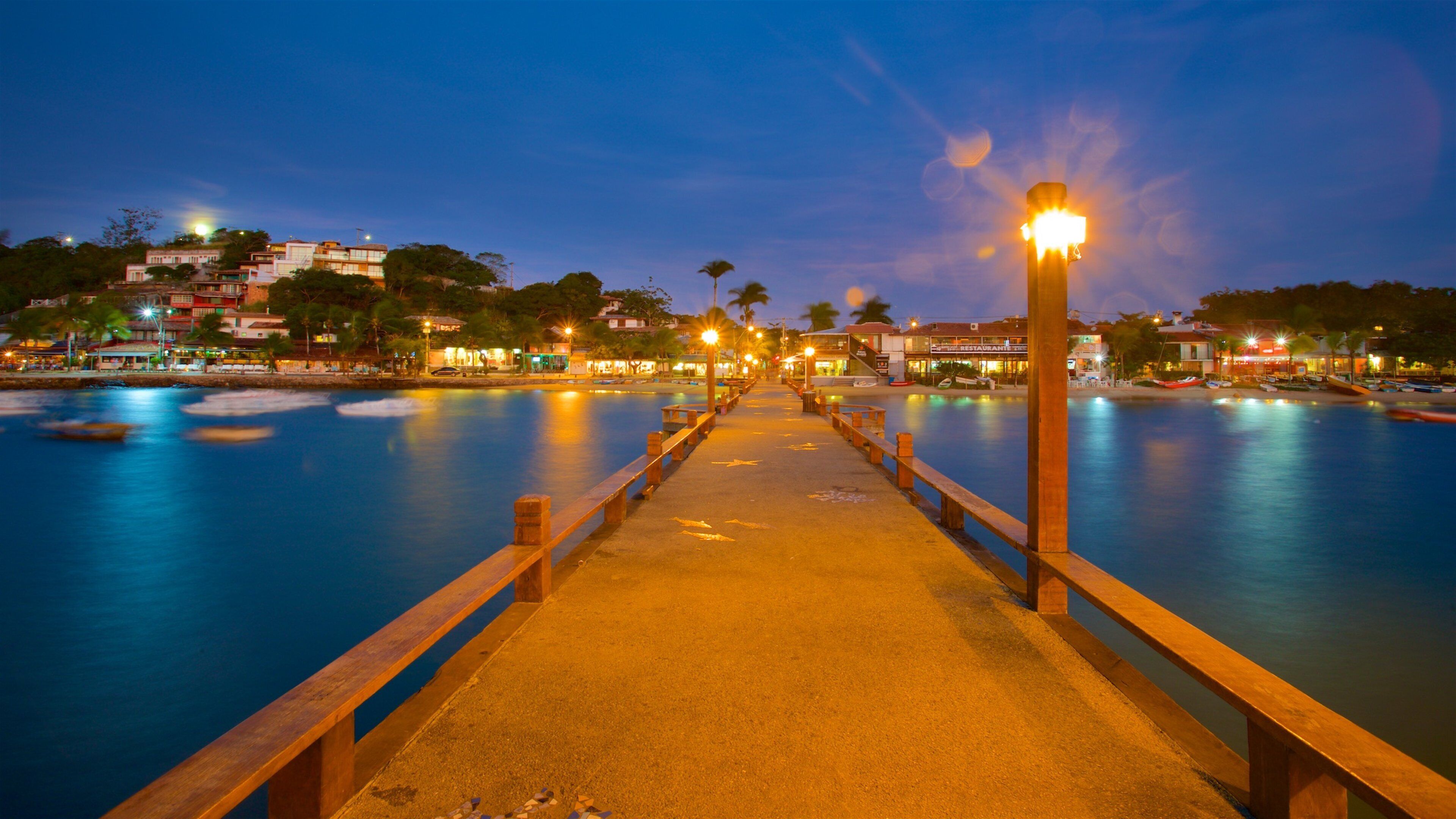 A waterfront promenade at dusk with a wooden walkway leading to a pool, surrounded by buildings and palm trees.