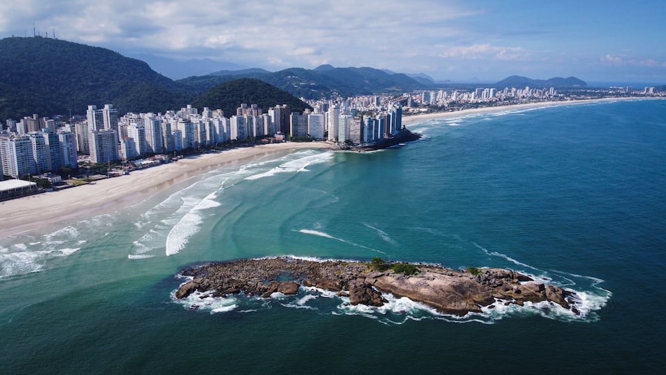 A coastal cityscape with a beach, high-rise buildings, and a rocky outcrop in the water.