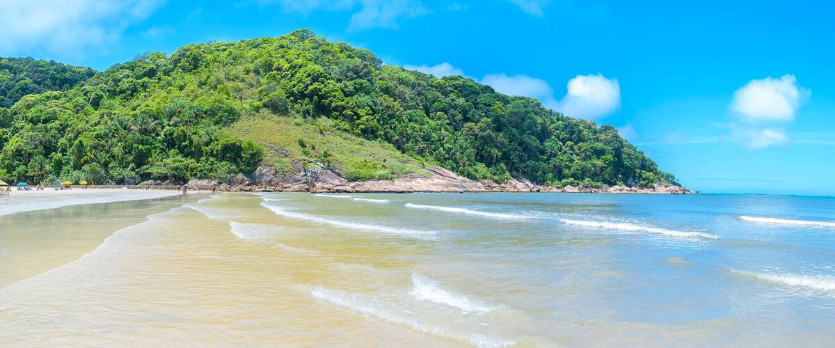 A beach with clear turquoise waters, a sandy shore, and a lush green hill in the background.