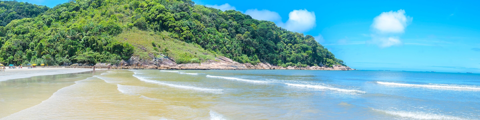 A beach with clear turquoise waters, a sandy shore, and a lush green hill in the background.