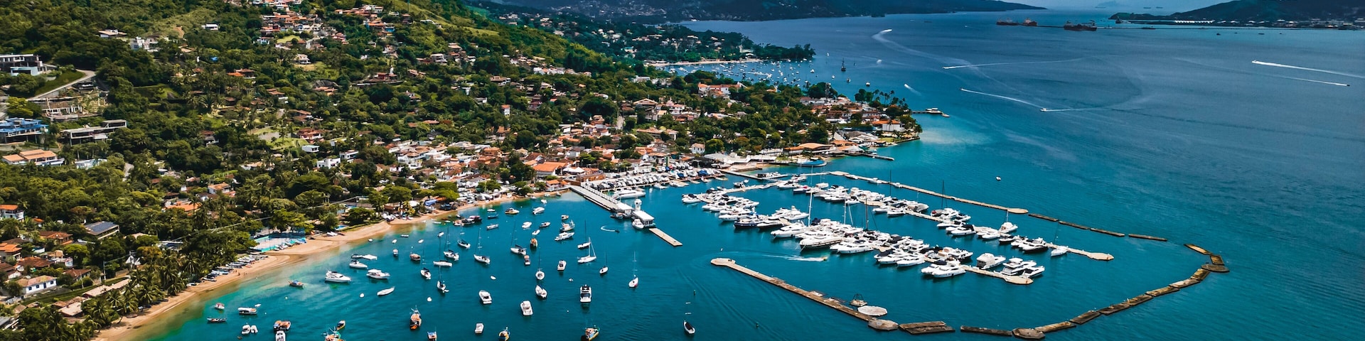 Uma cidade costeira com um porto, barcos e montanhas ao fundo.