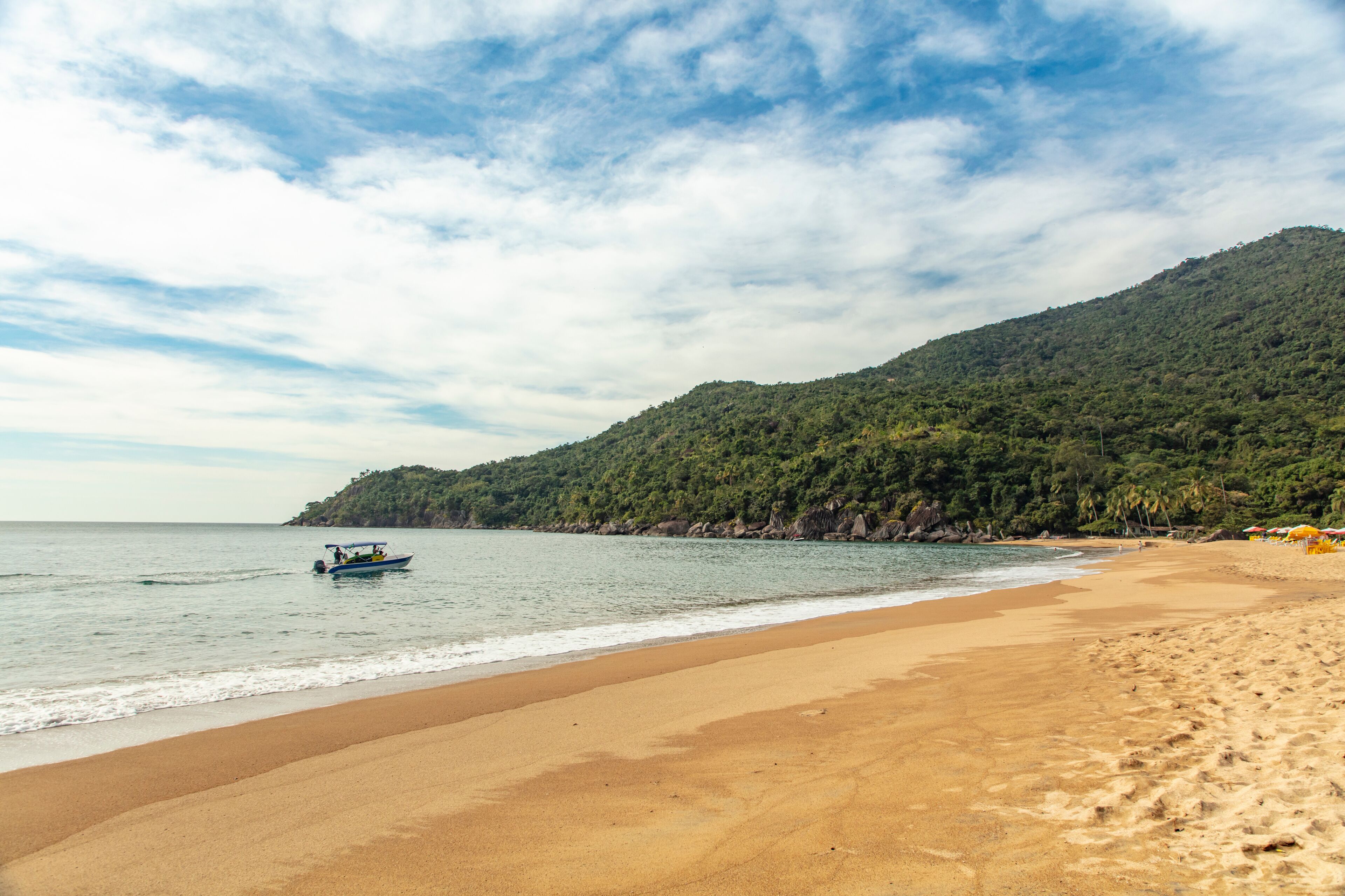 A beach with a boat on the water, a sandy shore, and a forested hill in the background.