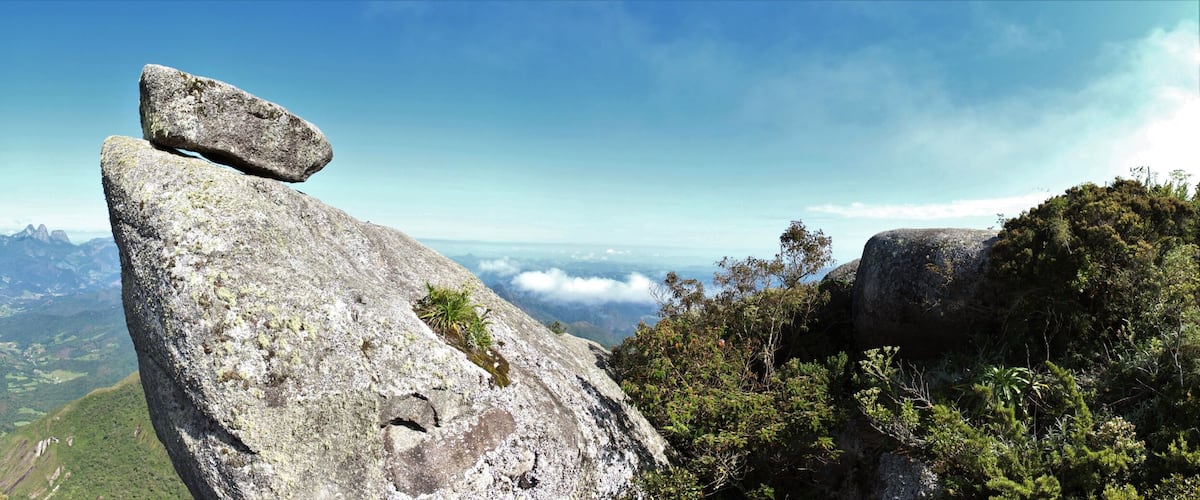 A prominent rock formation with a balanced boulder atop, surrounded by lush green forest and distant mountain ranges under a clear sky.