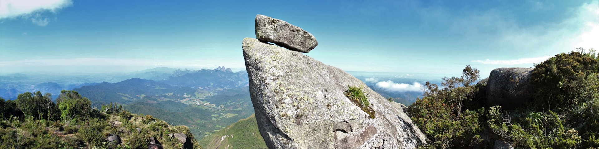 A prominent rock formation with a balanced boulder atop, surrounded by lush green forest and distant mountain ranges under a clear sky.