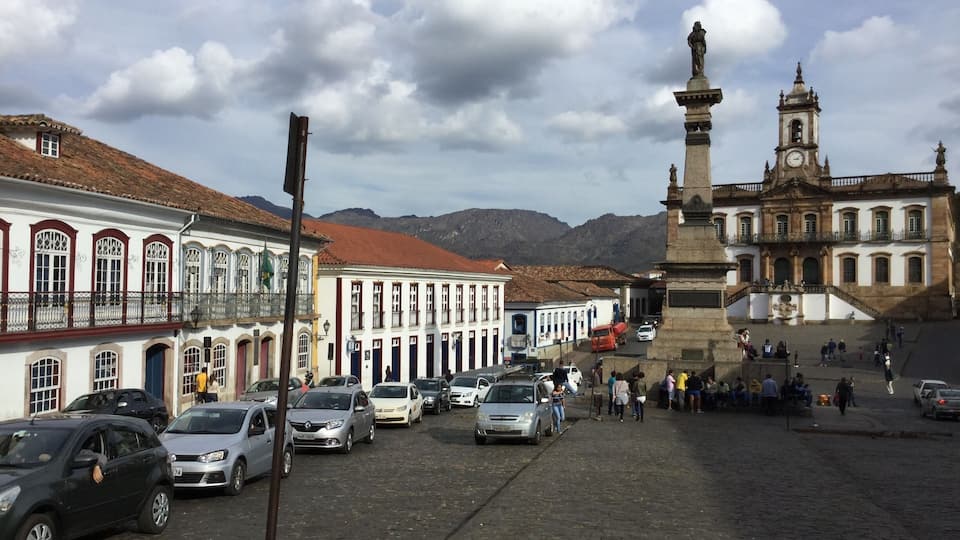 Una calle empedrada flanqueada por edificios históricos, una destacada torre del reloj y una estatua en una plaza.