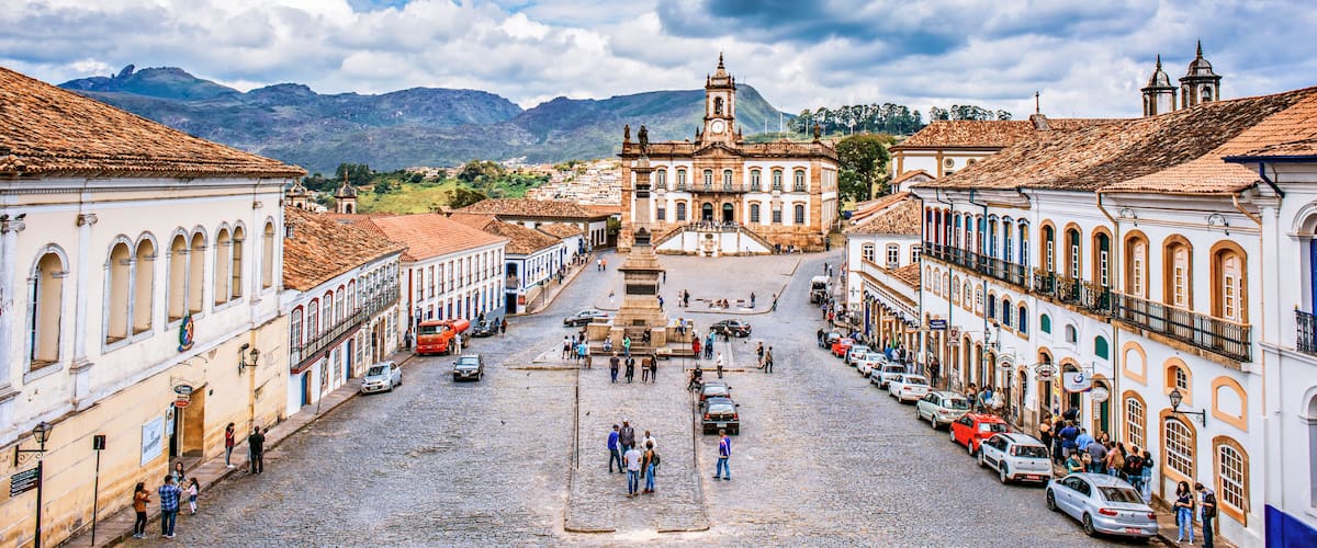 A cobblestone square with historic buildings, a prominent church with a clock tower, and mountains in the background.
