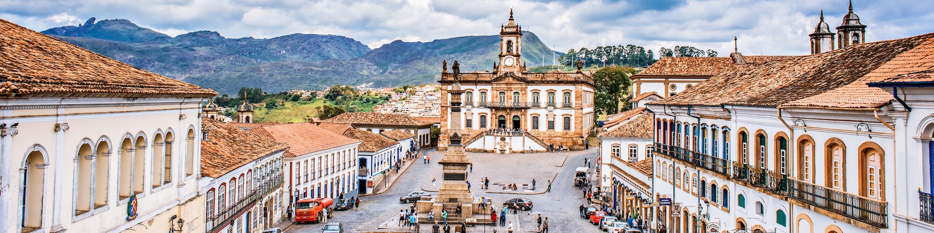 A cobblestone square with historic buildings, a prominent church with a clock tower, and mountains in the background.