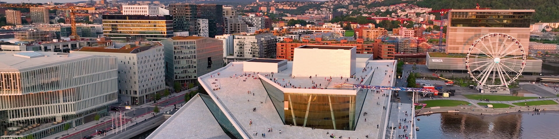 Um complexo arquitetônico moderno com uma grande fachada de vidro, um calçadão à beira-mar e uma roda-gigante ao fundo.