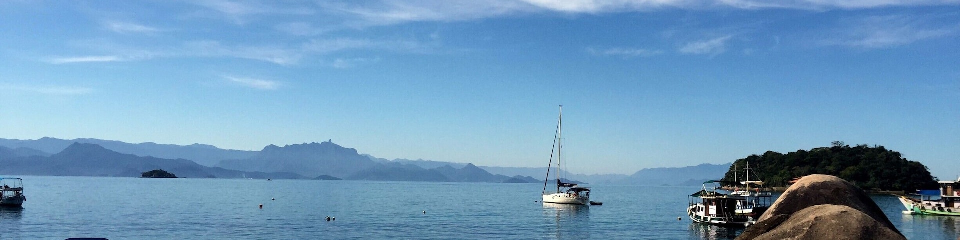 Une plage avec des bateaux, un voilier et des montagnes à l’horizon.