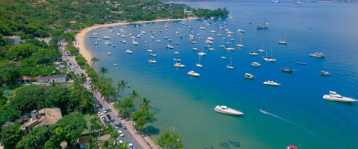 A coastal town with a marina filled with boats, surrounded by lush greenery and mountains in the background.