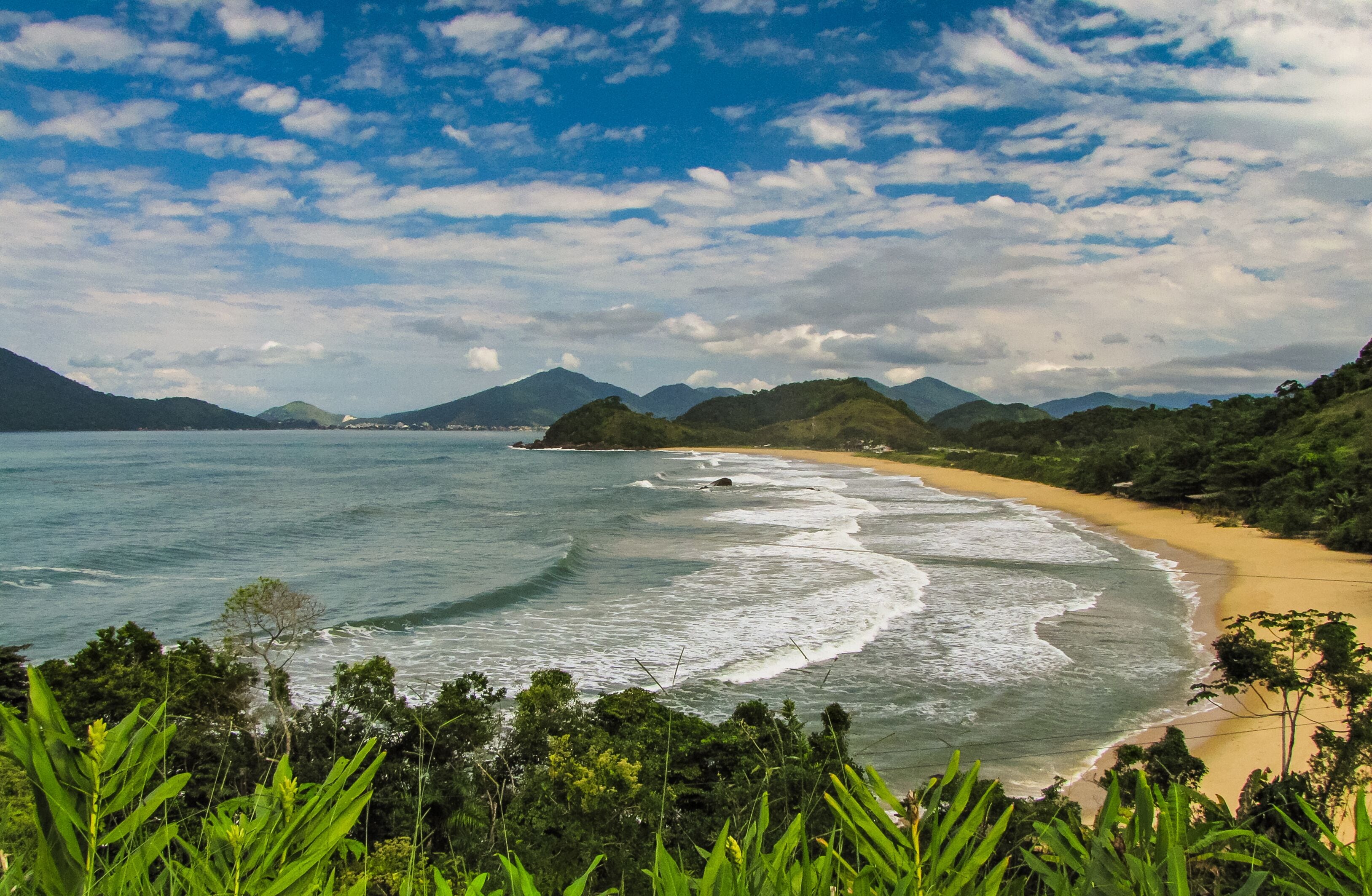 Panoramic view from a lookout of Praia Vermelha do Norte, Ubatuba/SP, Brazil. Sunny day with turquoise waves, preserved nature, and a clear sky dotted with white clouds.