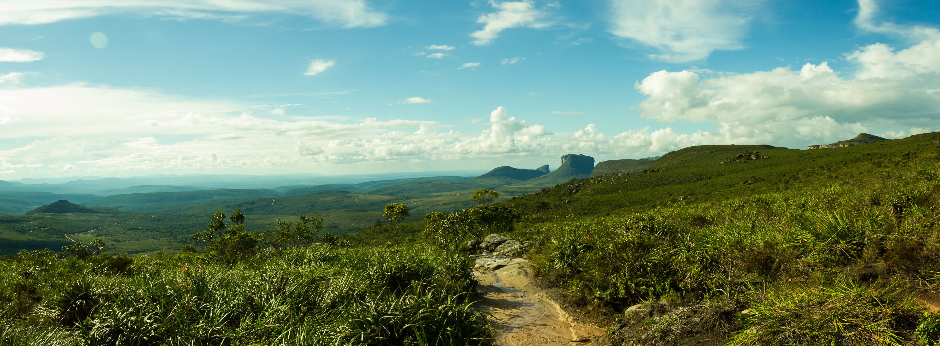 A hiking trail through a lush green landscape with rolling hills and distant mountains under a partly cloudy sky.